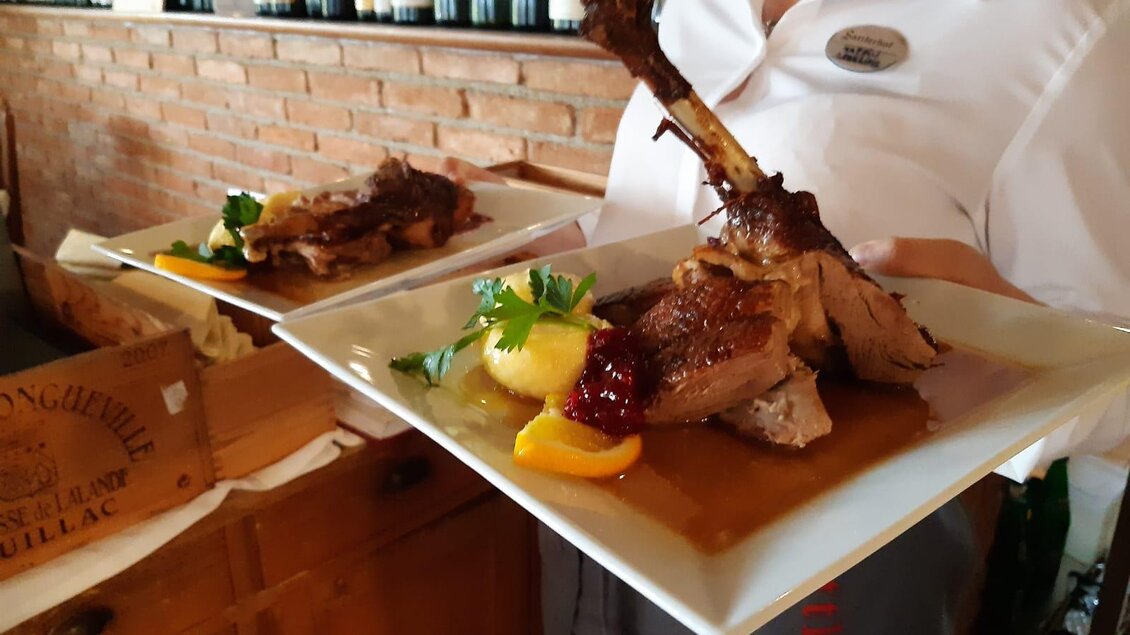 Ein Kellner serviert zwei Teller mit gebratenem Fleisch, garniert mit Fruchtstücken und einer Soße. Im Hintergrund sind Weinflaschen sichtbar. | © Sattler