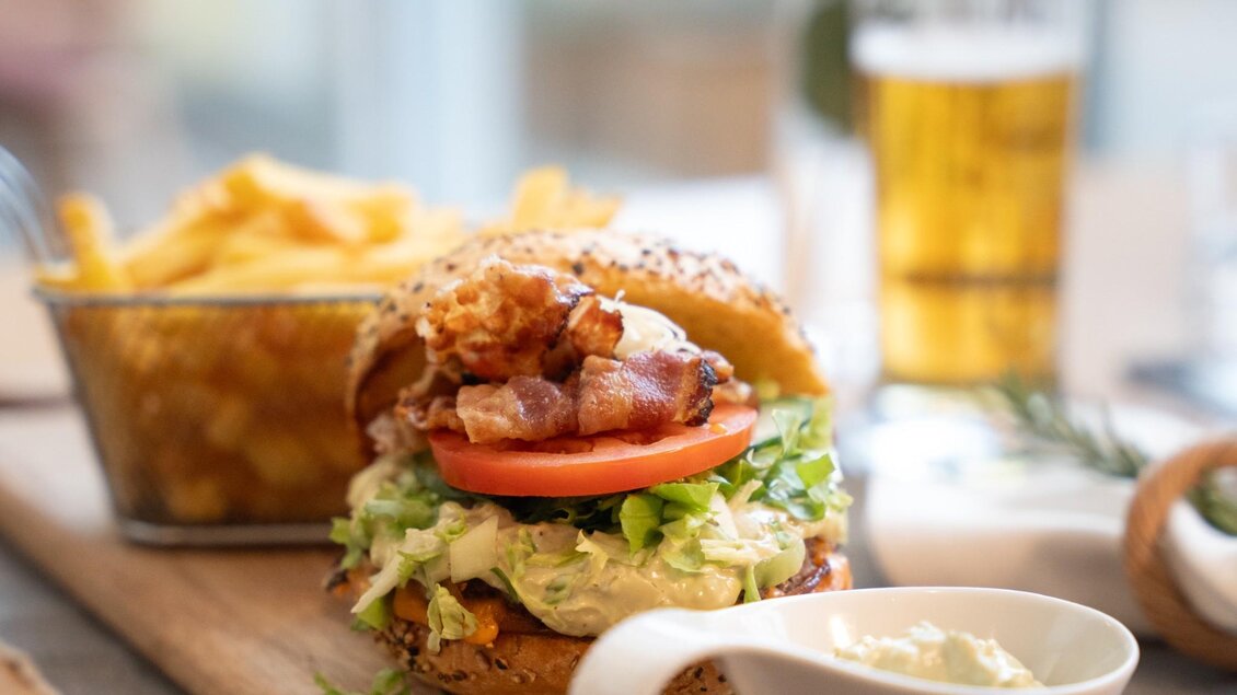 Burger mit Gemüse und Speck, dazu Pommes und ein Bier auf einem Holztisch im Schachenwald | © Schachenwald - Wolfgang Hummer