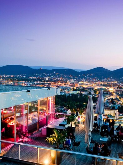 A rooftop bar with a spectacular view of the city and the surrounding mountains at dusk. The city lights sparkle beneath a colorful sky. | © Schlossberg - Werner Krug