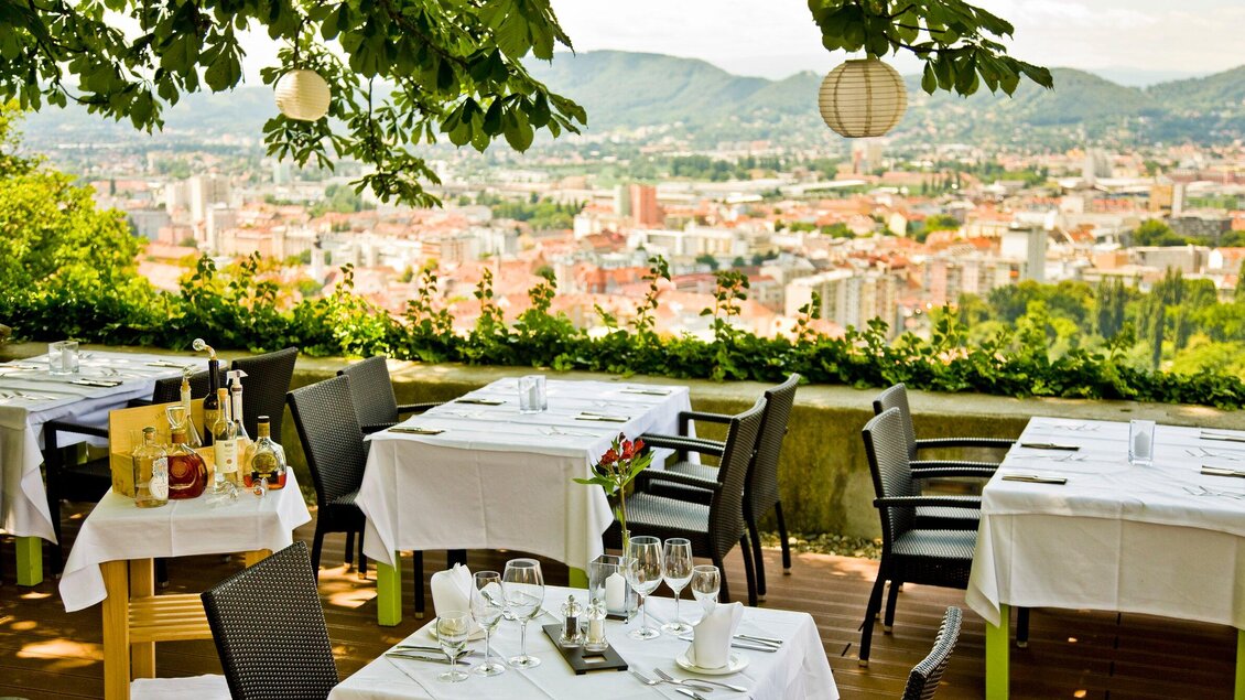 Ein elegantes Restaurant im Freien mit Blick auf die Stadt. Die Tische sind festlich gedeckt und von Bäumen umgeben. | © Schlossberg Restaurant - Werner Krug