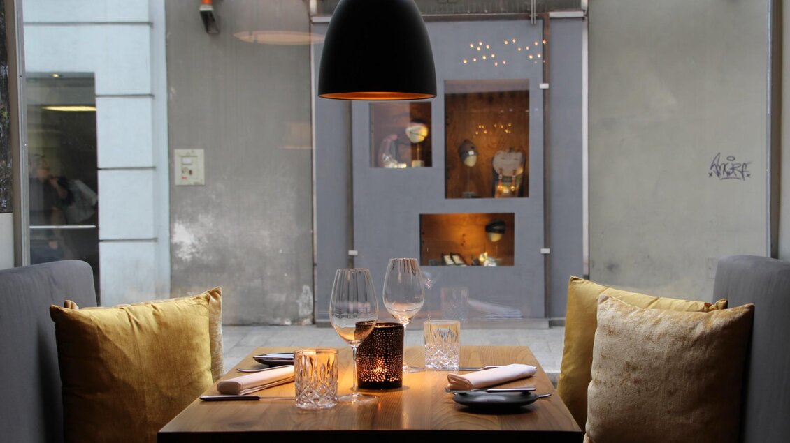 Ein elegantes Restaurant mit einem holzfarbenen Tisch und gemütlichen Sitzkissen. Der Tisch ist für zwei Personen gedeckt und eine moderne Lampe hängt darüber. | © Schmidhofer im Palais