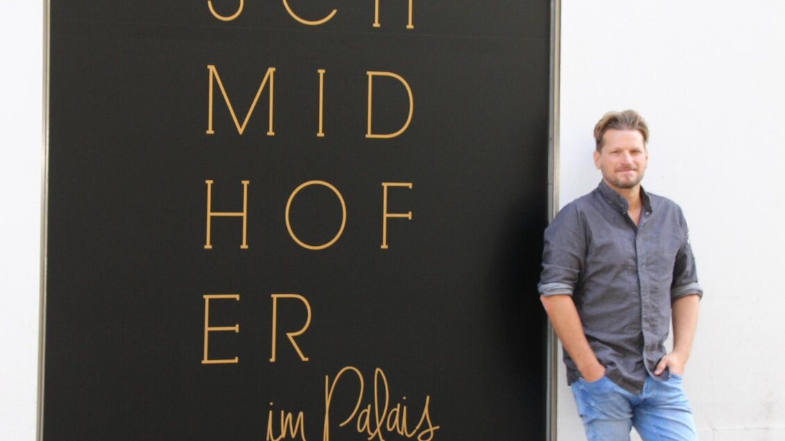 Ein Mann steht neben einem großen Schild mit dem Schriftzug "SCHMIDHOFER". Der Hintergrund ist schlicht und bietet einen klaren Kontrast zu den goldenen Buchstaben. | © Restaurant Schmidhofer