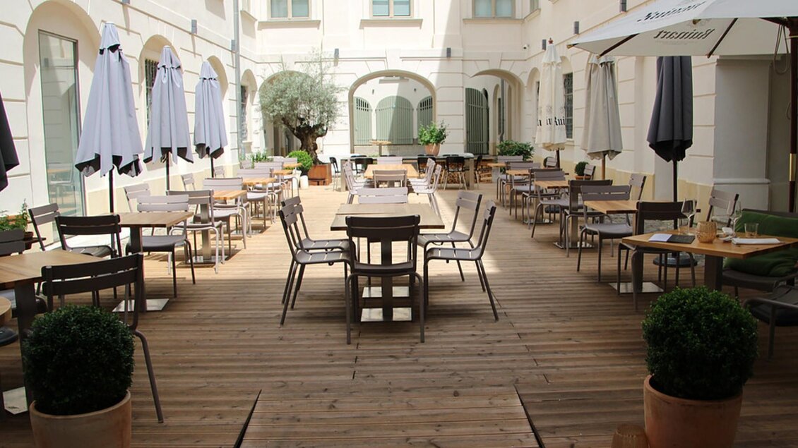 Ein ruhiger Innenhof mit Holzfußboden und mehreren Tischen. Schirme spenden Schatten, umgeben von Pflanzen. | © Schmidhofer im Palais