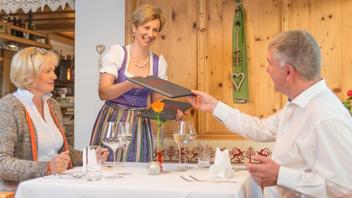 Eine Kellnerin in traditioneller Tracht serviert einem Paar am Tisch ein Menü. Der Tisch ist festlich gedeckt mit Gläsern und einer Blume. | © Restaurant Schwaiger