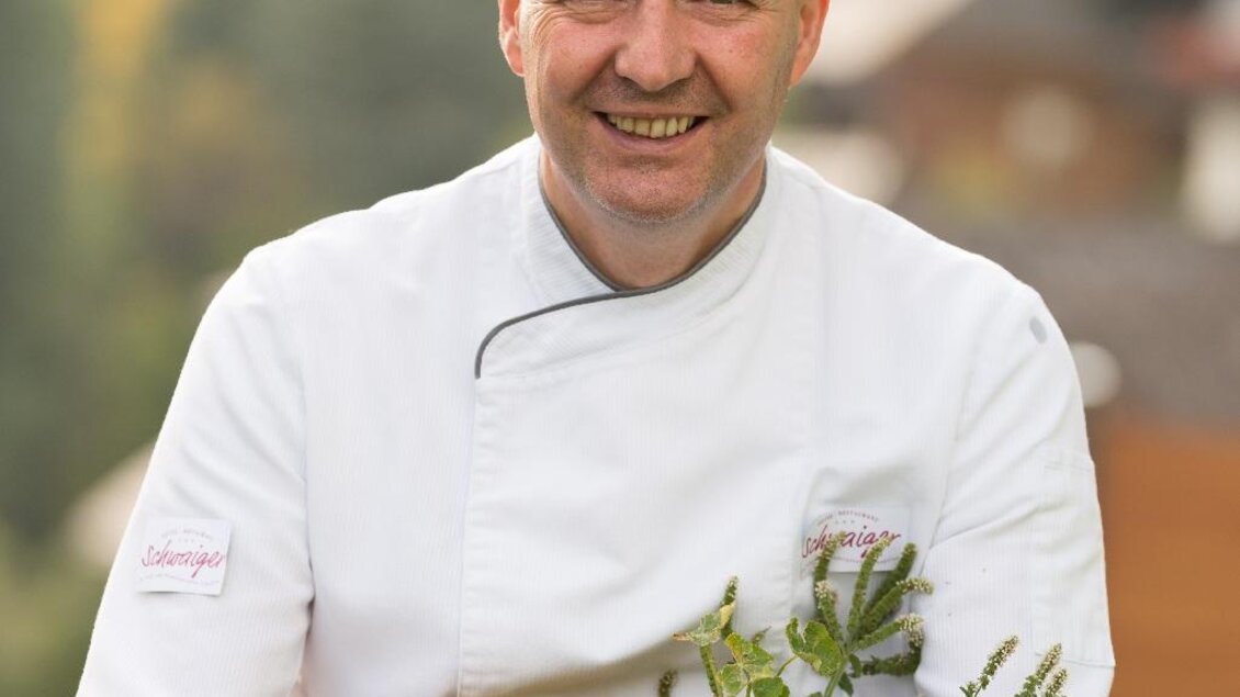 Ein lächelnder Koch hält einen Korb mit frischen Kräutern und essbaren Blumen. Im Hintergrund sind grüne Bäume und eine ländliche Umgebung zu sehen. | © Restaurant Schwaiger