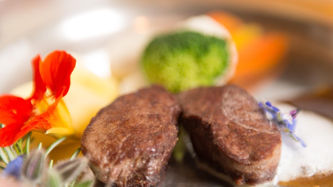 Zwei saftige Fleischstücke auf einem eleganten Teller, begleitet von buntem Gemüse und essbaren Blüten. Das Gericht vermittelt eine ansprechende Präsentation und kreative Kochkunst. | © Restaurant Schwaiger