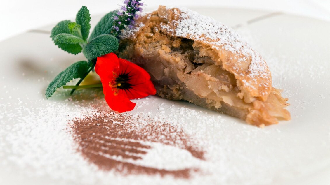 Ein Stück Apfelstrudel auf einem weißen Teller, dekoriert mit Puderzucker und essbaren Blumen. Die Füllung ist sichtbar und sieht köstlich aus. | © Restaurant Schwaiger