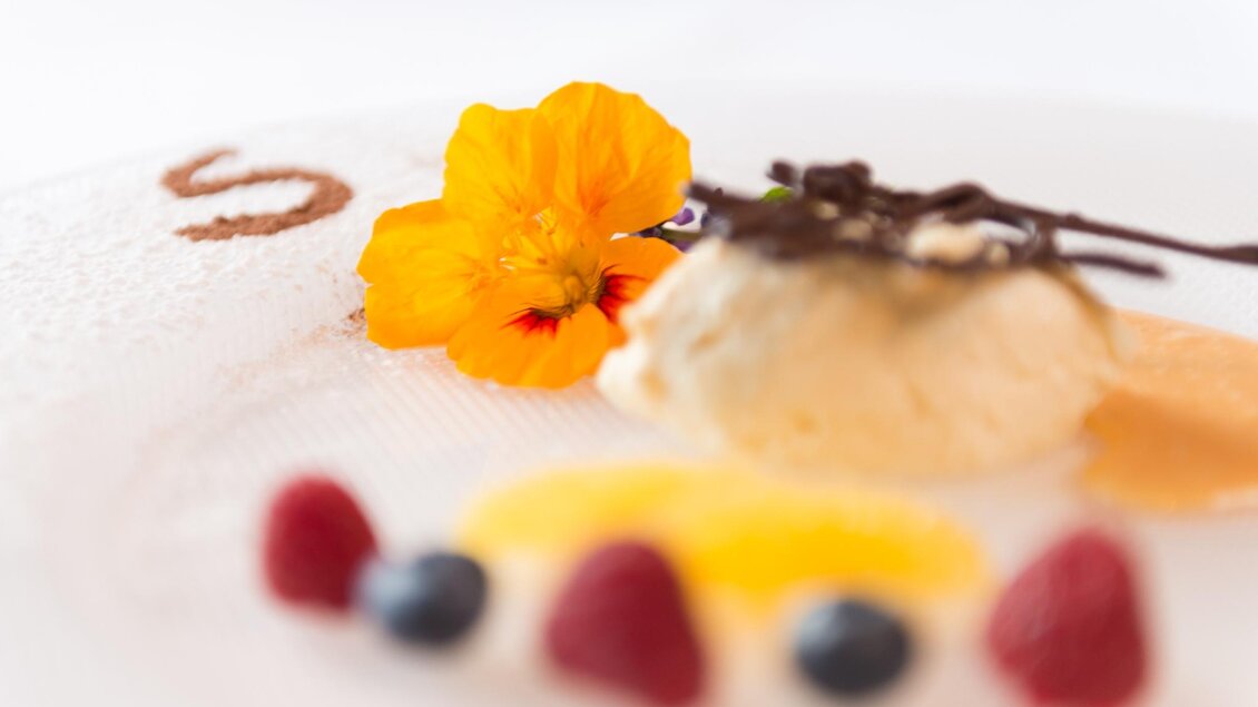 Ein kunstvolles Dessert auf einem Teller, dekoriert mit einer Blume und unterschiedlichen Früchten. Das Gericht zeigt eine Kombination aus Eiscreme und Schokoladendekoration. | © Restaurant Schwaiger