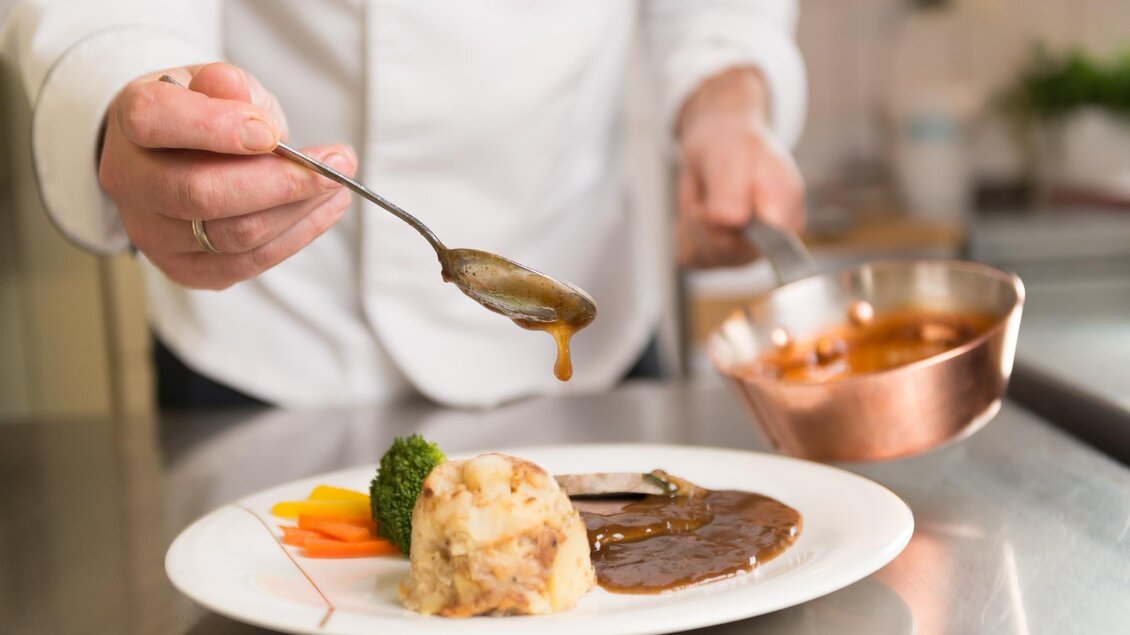 Ein Koch reicht eine Portion Fleisch mit Soße auf einem Teller. Daneben sind Gemüse und ein Kartoffelbrei dekorativ angerichtet. | © Restaurant Schwaiger