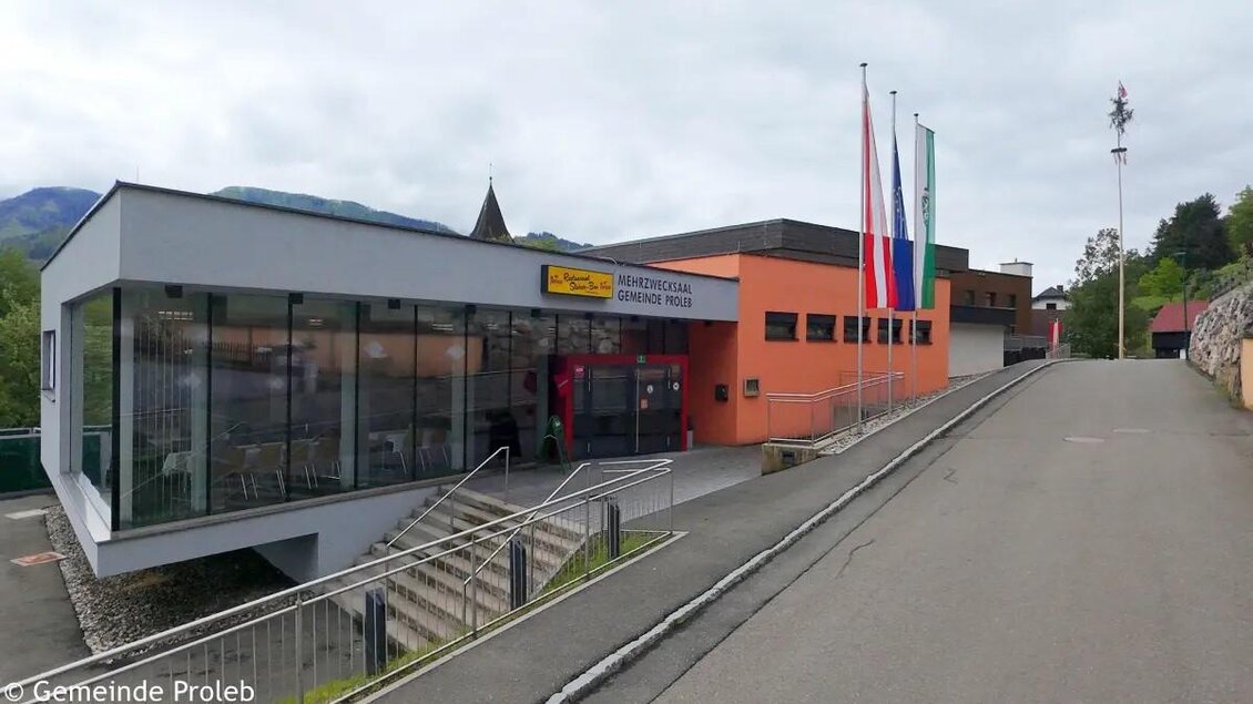 Ein modernes Gebäude mit großen Fenstern und mehreren Fahnen vor dem Eingang. Die umliegende Straße führt sanft bergauf. | © Gemeinde Proleb