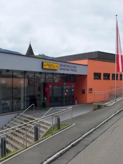 Ein modernes Gebäude mit großen Fenstern und mehreren Fahnen vor dem Eingang. Die umliegende Straße führt sanft bergauf. | © Gemeinde Proleb