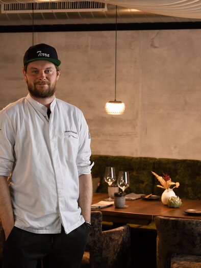 A chef stands in a stylish restaurant in front of a fireplace. The decor is modern and cozy, with elegant tables and chairs. | © achromaticphotography