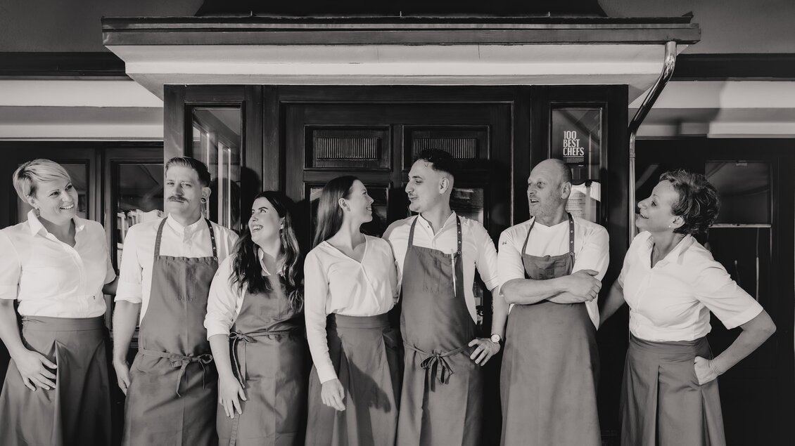 Eine Gruppe von sieben Personen steht vor einer Tür, alle tragen ähnliche Kittelschürzen. Sie wirken freundlich und lächeln in die Kamera. | © Die Abbilderei