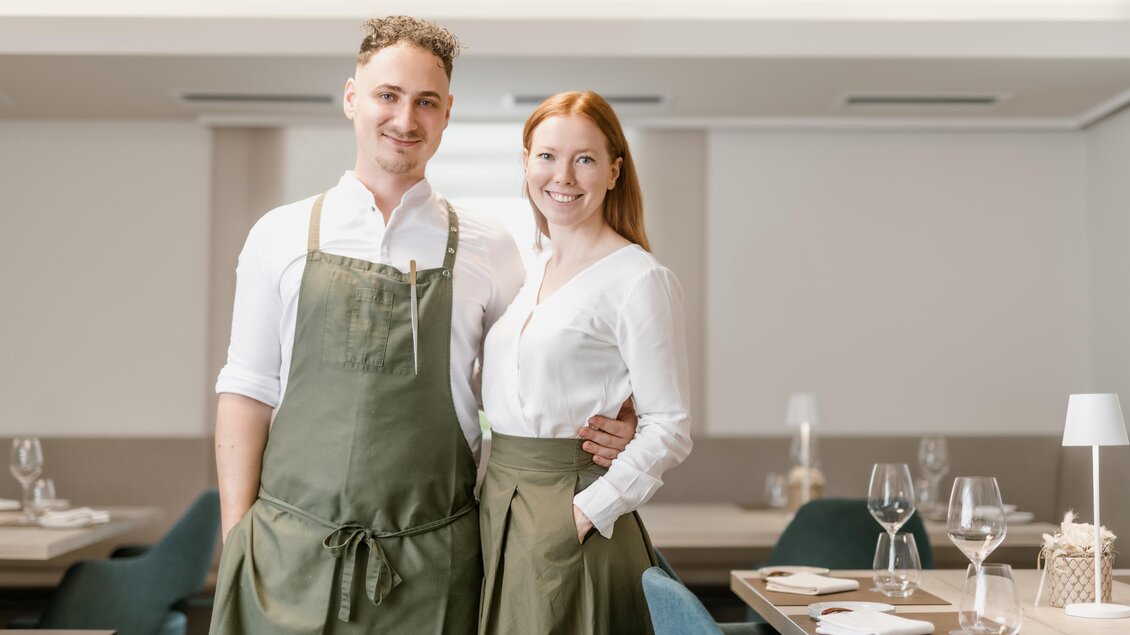 Ein Paar steht in einem eleganten Restaurant. Sie tragen Arbeitskleidung und lächeln freundlich in die Kamera. | © Die Abbilderei