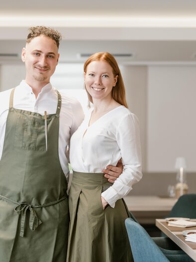 Ein Paar steht in einem eleganten Restaurant. Sie tragen Arbeitskleidung und lächeln freundlich in die Kamera. | © Die Abbilderei