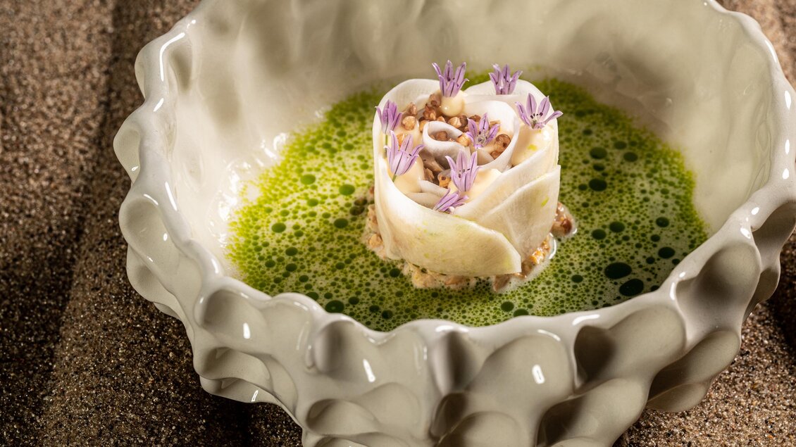 Ein kunstvoll angerichtetes Gericht in einer eleganten Schüssel. Die Speise besteht aus einer zarten Rolle und ist mit frischen Kräutern und einer grünen Sauce umgeben. | © Die Abbilderei