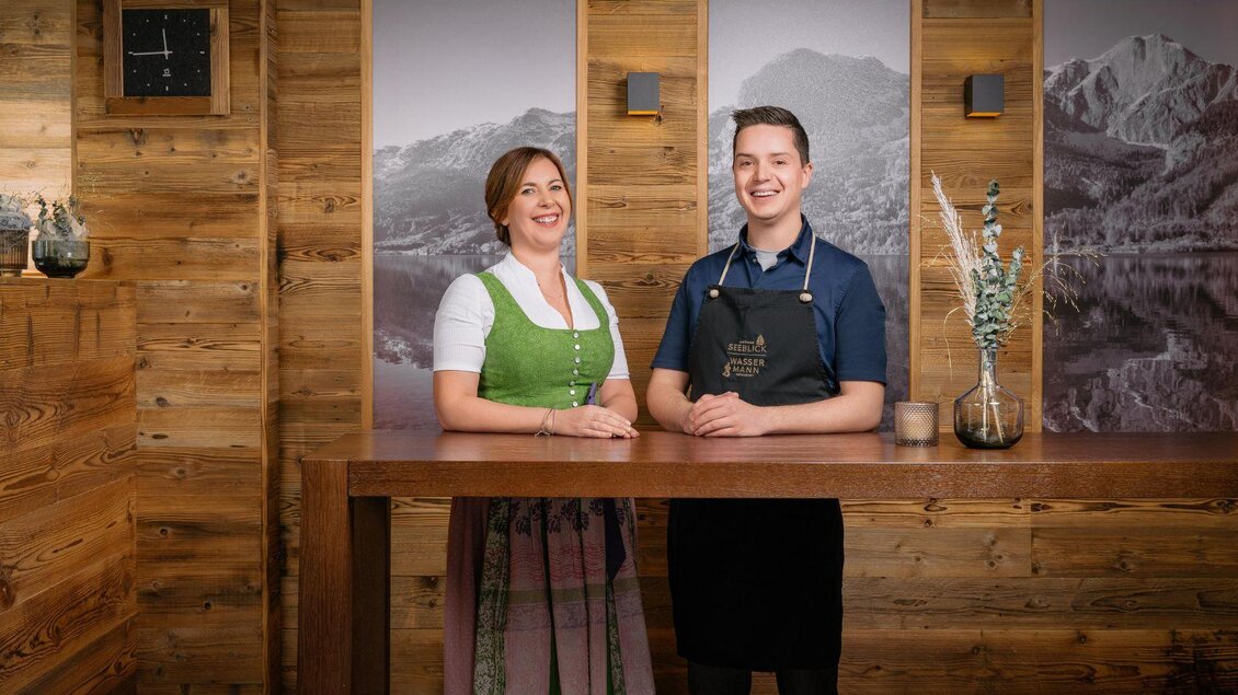 Zwei freundliche Mitarbeiter stehen an einem Holztresen. Im Hintergrund sind Landschaftsbilder mit Bergen zu sehen. | © Mondi Resort