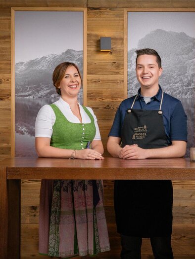 Two friendly employees stand at a wooden counter. In the background, landscape pictures featuring mountains can be seen. | © Mondi Resort