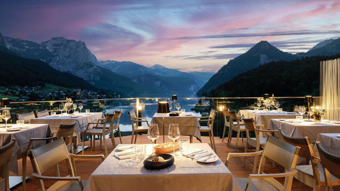 Ein elegantes Restaurant mit Tischen auf einer Terrasse. Im Hintergrund sind Berge und ein schöner Sonnenuntergang zu sehen. | © Mondi Resort