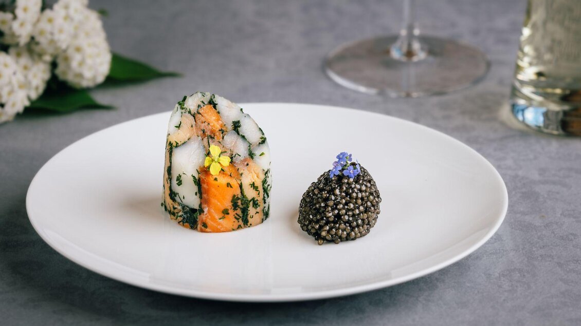 Ein elegantes Gericht auf einem weißen Teller. Es besteht aus einer bunte Fischpraline und einer kleinen Portion Kaviar, garniert mit Blumen. | © Mondi Resort