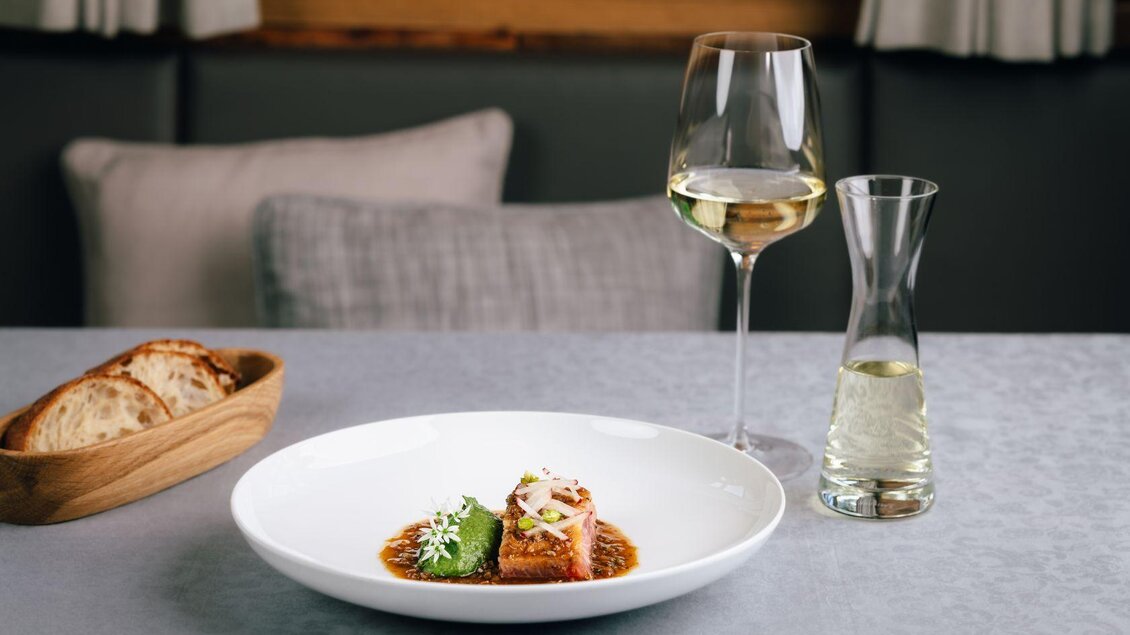 Ein elegantes Gericht auf einem weißen Teller, garniert mit frischen Kräutern. Dazu ein Glas Weißwein und ein Krug mit einem weiteren Getränk, sowie frisches Brot in einer Holzschale. | © Mondi Resort