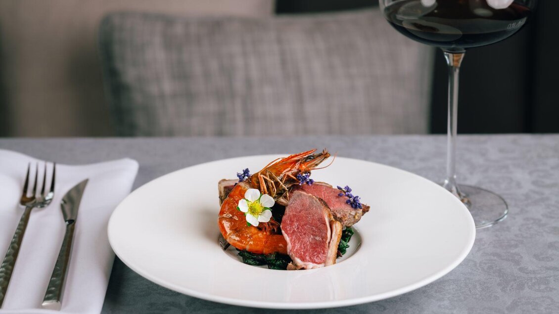Ein elegantes Gericht mit zartem Fleisch, Garnelen und essbaren Blüten. Dazu ein Glas Rotwein auf einem stilvollen Tisch. | © Mondi Resort