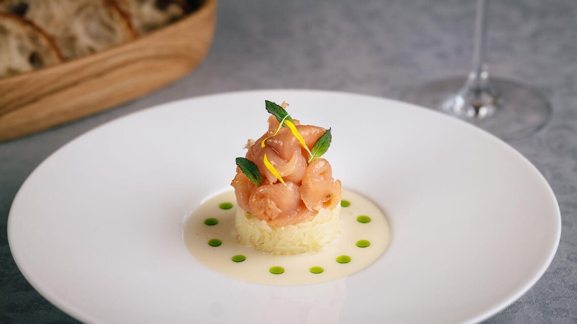 Ein stilvolles Gericht auf einem weißen Teller, bestehend aus Garnelen und Kräutern. Umgeben von einer feinen Sauce und dekorierten Punkten. | © Mondi Resort