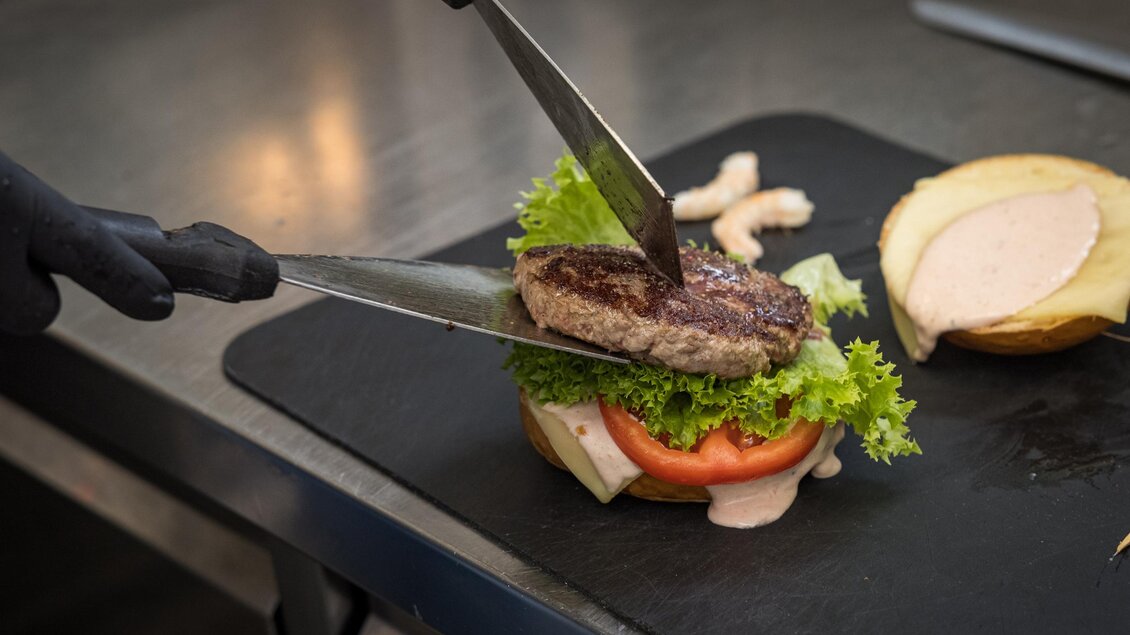 Ein frisch zubereiteter Burger wird mit einem Patties, Salat und Tomate geschichtet. Die Zutaten liegen auf einem Schneidebrett in einer modernen Küche. | © Netzwerk Kulinarik wildbild