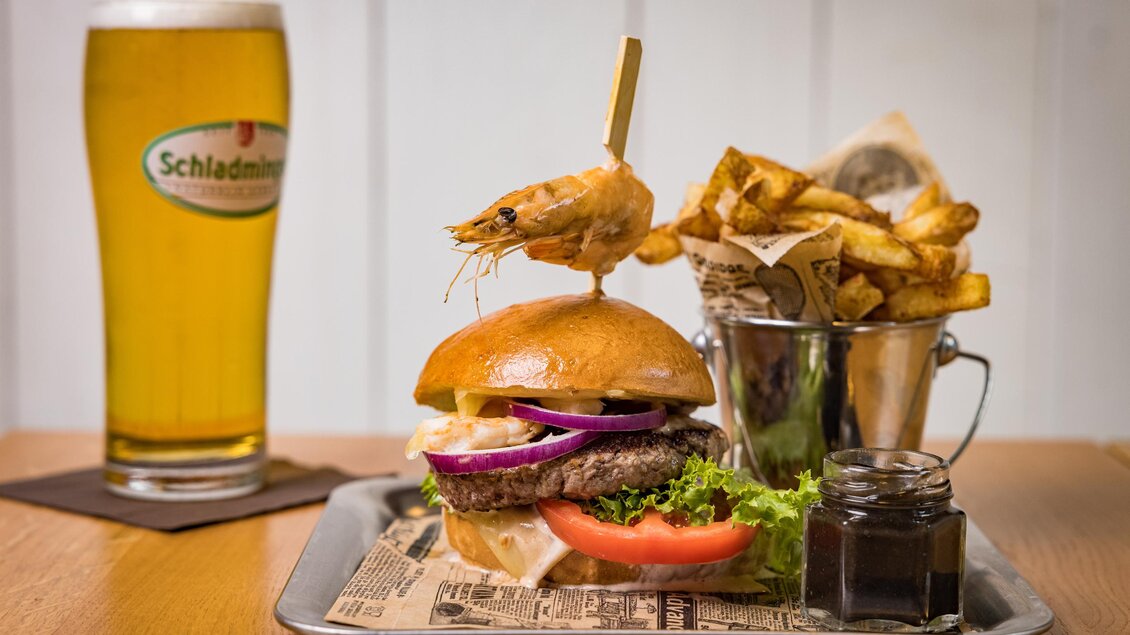 Ein köstlicher Burger mit Rindfleisch, frischem Gemüse und einer Garnele als Dekoration. Dazu gibt es knusprige Pommes frites und ein Glas Bier. | © Netzwerk Kulinarik wildbild