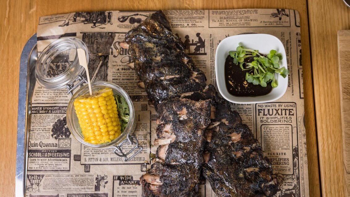 Saftiges gegrilltes Fleisch auf einer alten Zeitungsunterlage, serviert mit einer Portion Mais und einer kleinen Schale mit Soße. Das Gericht ist ansprechend angerichtet und lädt zum Essen ein. | © Netzwerk Kulinarik wildbild