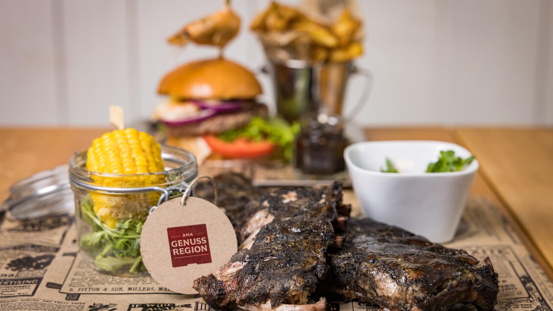 Ein Teller mit gegrillten Rippchen, frischem Salat und Maiskolben. Im Hintergrund sind ein Burger und Pommes frites zu sehen. | © Netzwerk Kulinarik wildbild