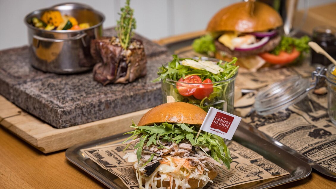 Ein köstlicher Burger mit frischem Salat und einer Tomate. Im Hintergrund befindet sich ein weiteres Gericht in einer Schale. | © Netzwerk Kulinarik wildbild