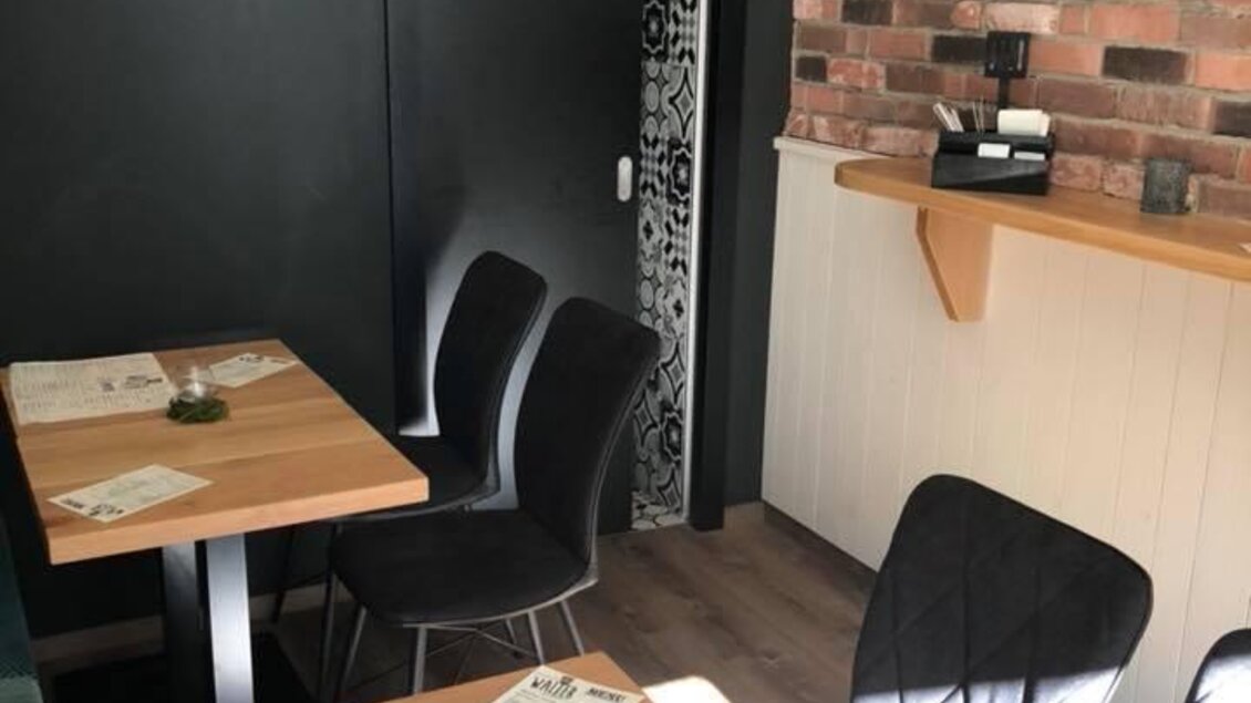 Ein modernes Restaurant mit Holzstühlen und großen Fenstern. Die Wände sind in dunklen Farben gehalten, und es gibt einen separaten Bereich mit einer Bar. | © Walter.Grill.Burger