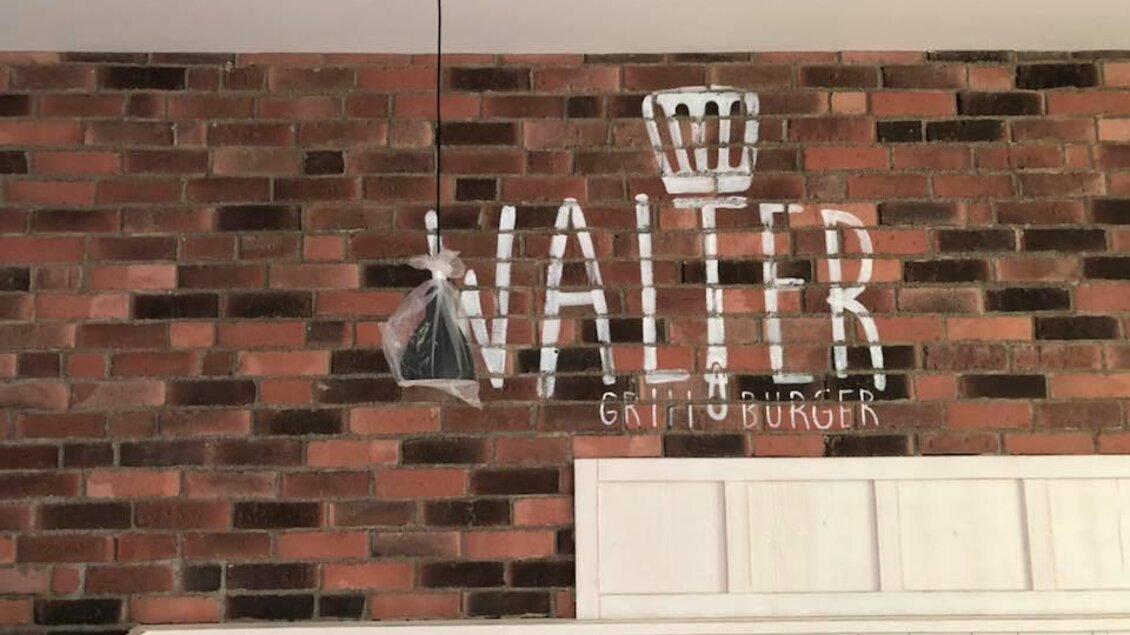 Eine rustikale Wand mit Backsteinoptik und der Schriftzug "WALTER GRILL BURGER". Darunter befindet sich eine weiße Holzverkleidung. | © Walter.Grill.Burger