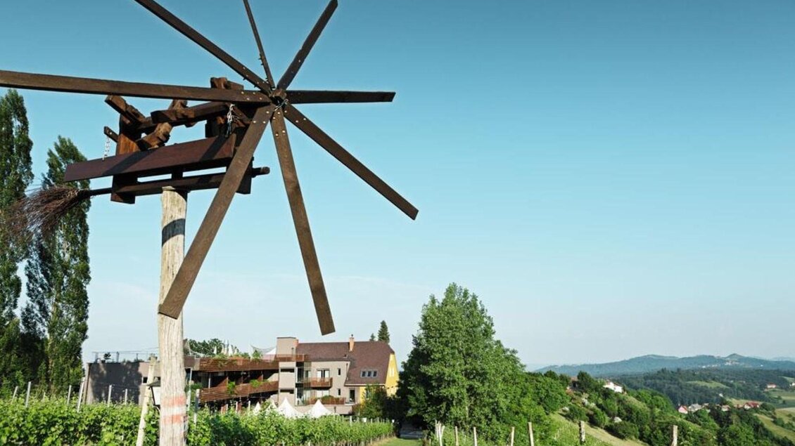 Ein malerischer Weinberg mit einem alten Windrad im Vordergrund. Im Hintergrund sieht man ein Haus und sanfte Hügel unter klarem Himmel. | © Weingartenhotel