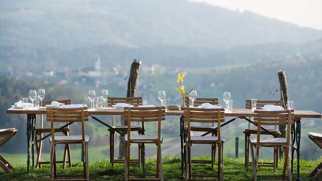 Ein schön gedeckter Tisch im Freien mit Blick auf eine hügelige Landschaft. Die Stühle sind aus Holz und die Landschaft im Hintergrund ist sanft und malerisch. | © Weingartenhotel