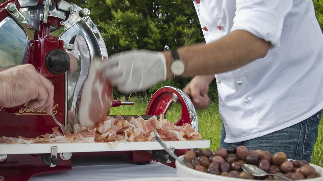 Ein Koch schneidet Schinken mit einer Schneidemaschine im Freien. Auf dem Tisch liegen frische Tomaten und andere Zutaten. | © Weingartenhotel