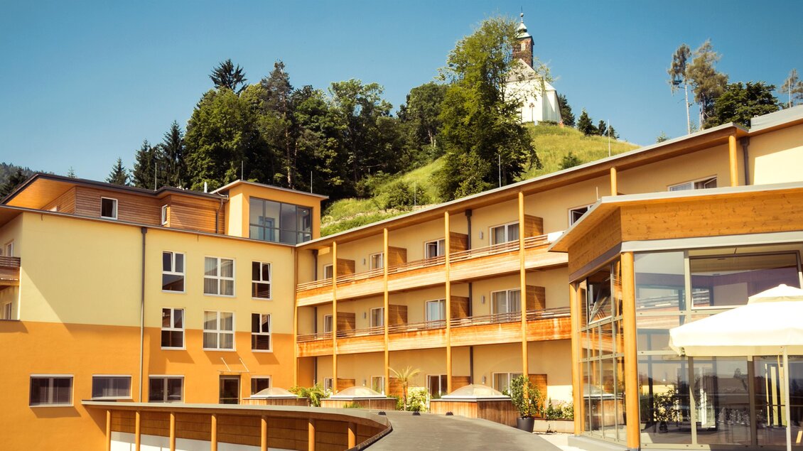 Ein modernes Hotelgebäude mit mehreren Etagen und großen Fenstern. Im Hintergrund ist ein Hügel mit einer kleinen Kapelle zu sehen. | © Lupi_Spuma