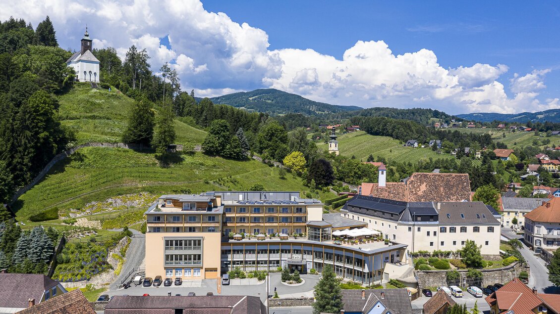 Eine malerische Landschaft mit einem modernen Hotel im Vordergrund. Im Hintergrund sind sanfte Hügel und eine kleine Kirche zu sehen.