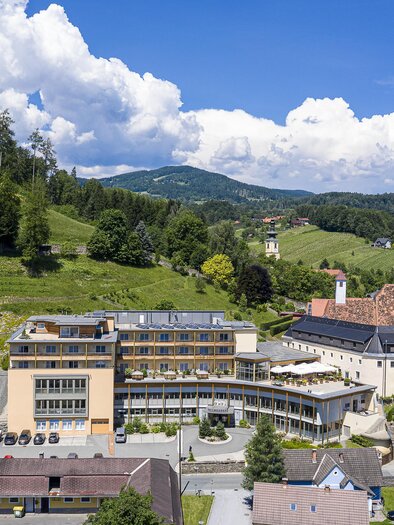 Eine malerische Landschaft mit einem modernen Hotel im Vordergrund. Im Hintergrund sind sanfte Hügel und eine kleine Kirche zu sehen.