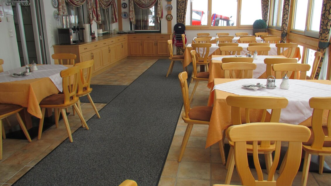 Ein gemütlicher Speisesaal mit orangefarbenen Tischdecken und Holzmöbeln. Das Zimmer hat große Fenster mit Blick nach draußen und bietet Platz für mehrere Gäste. | © Hotel Alpen Arnika
