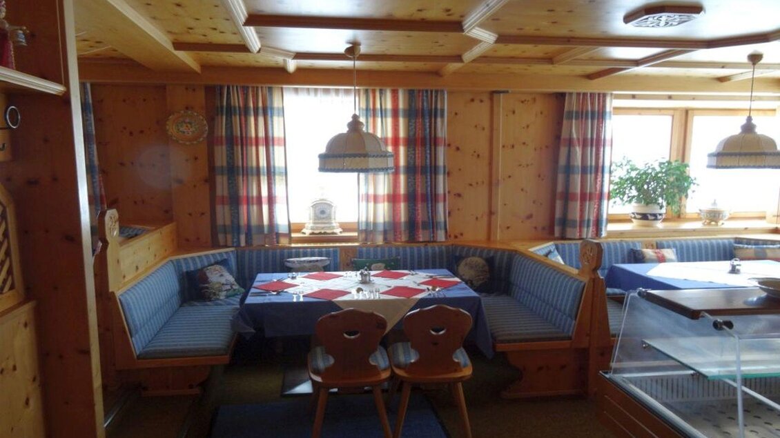 Ein gemütlicher Essbereich mit Holzwänden und blauen Sitzbänken. Der Tisch ist dekoriert und die Fenster sind mit karierten Vorhängen geschmückt. | © Hotel Alpen Arnika