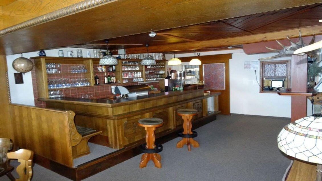 Eine gemütliche Bar mit Holzdetails und freundlicher Atmosphäre. Am Tresen stehen drei Barhocker, und im Hintergrund sind Regale mit Getränken sichtbar. | © Hotel Alpen Arnika