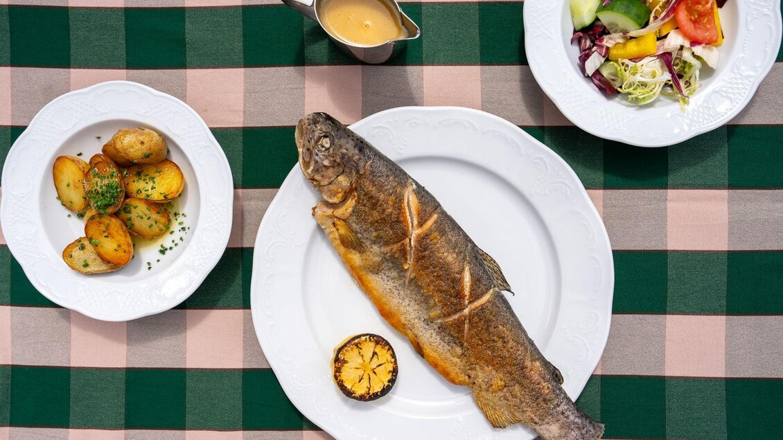 Ein gegrillter Fisch auf einem weißen Teller, dazu gebratene Kartoffeln und ein frischer Salat. Die Speisen sind auf einem karierten Tischcloth angerichtet. | © Hotel Alpen Arnika