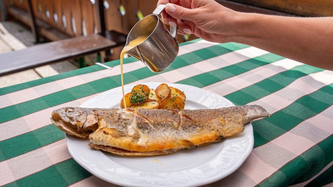 Ein frisch zubereiteter Fisch auf einem weißen Teller, serviert mit Kartoffeln und Gemüse. Dazu wird eine Sauce aus einer Kelle über den Fisch gegossen. | © Hotel Alpen Arnika