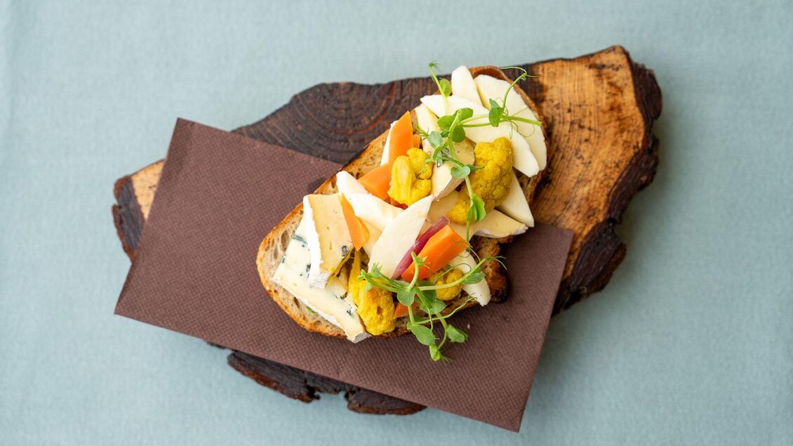 Ein rustikales Brot mit verschiedenen Gemüse- und Käsesorten, dekoriert mit frischen Kräutern. Es liegt auf einem Holzbrett und ist ansprechend angerichtet. | © Hotel Alpen Arnika