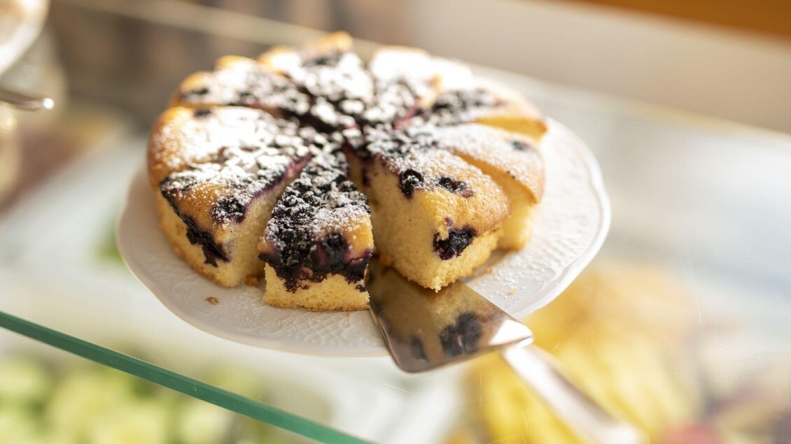 Ein geschnittener Kuchen auf einem weißen Teller, bestreut mit Puderzucker. Der Kuchen hat eine fruchtige Füllung und liegt auf einem Glastisch. | © Hotel Alpen Arnika
