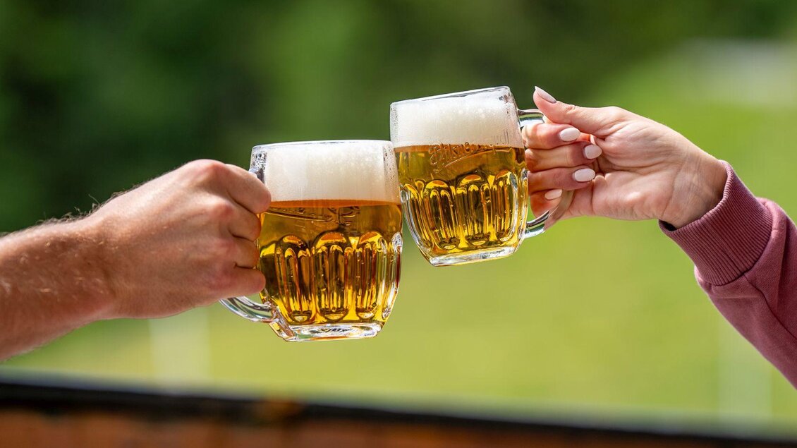 Zwei Bierkrüge stoßen aneinander. Im Hintergrund ist eine grüne Landschaft zu sehen. | © Hotel Alpen Arnika