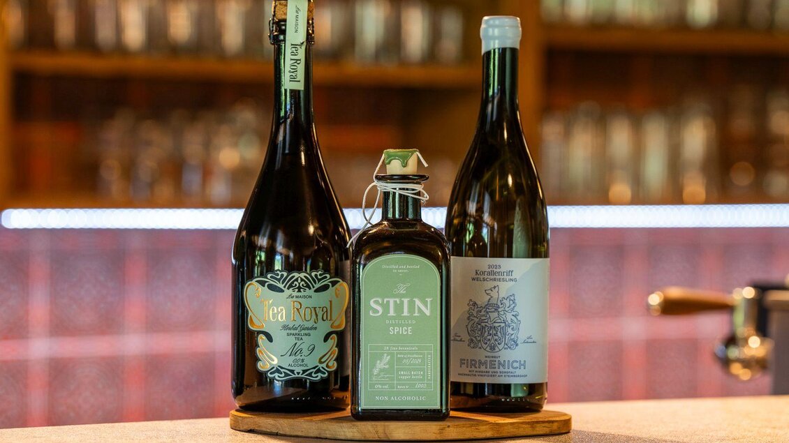 Drei Flaschen alkoholischer Getränke stehen auf einem Holzbrett. Im Hintergrund sind verschwommene Gläser und eine Bar zu sehen. | © Hotel Alpen Arnika