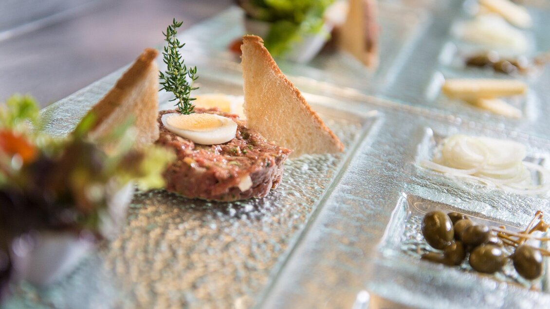 Ein kunstvoll angerichtetes Gericht mit Tatar, garniert mit einem kleinen Ei und frischen Kräutern. Daneben befinden sich verschiedene Beilagen und Zutaten auf einem gläsernen Teller. | © Ulf Thausing Photography